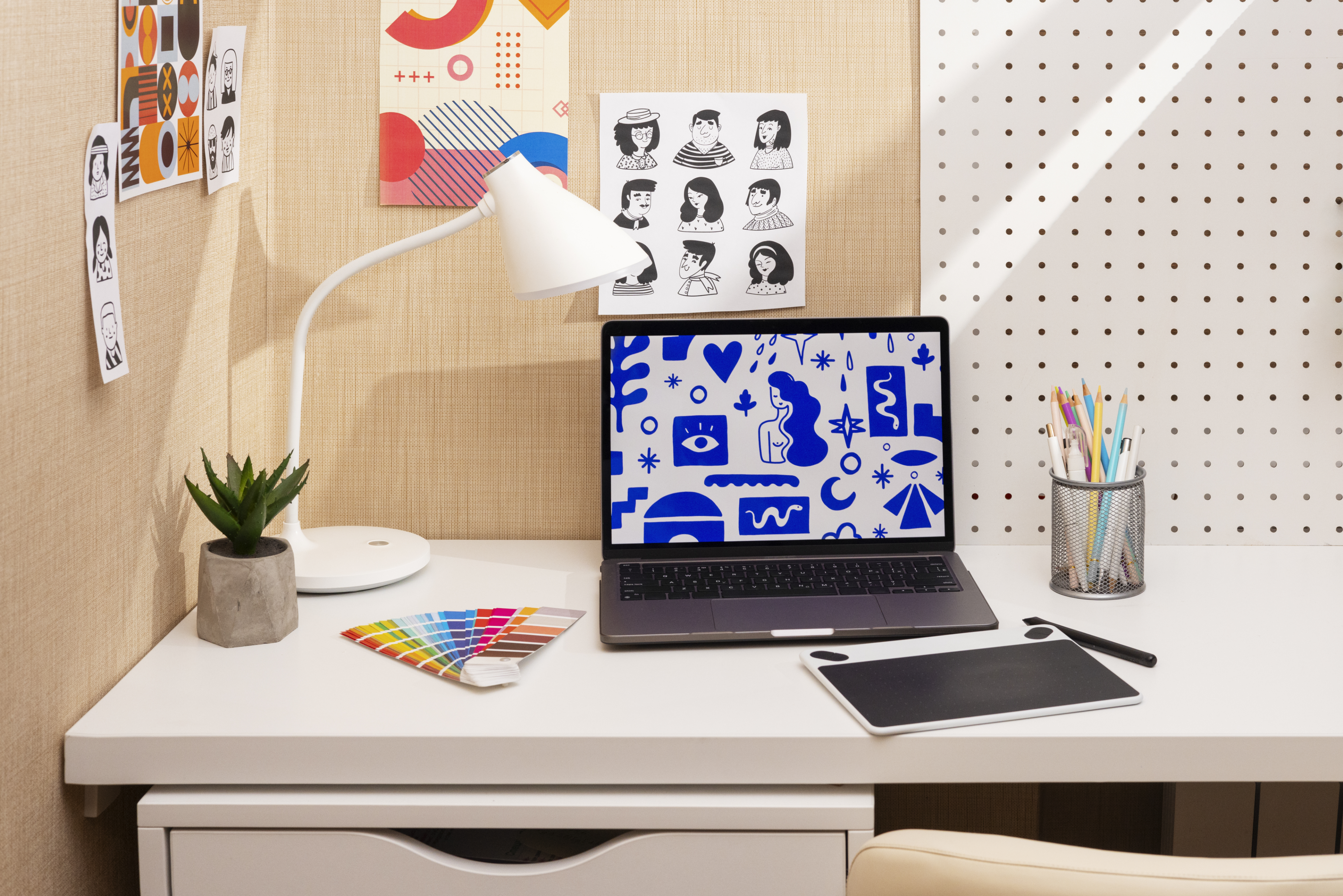 Designer's desk with a laptop showing brand illustration work, a color swatch fan, a graphics tablet and stationery