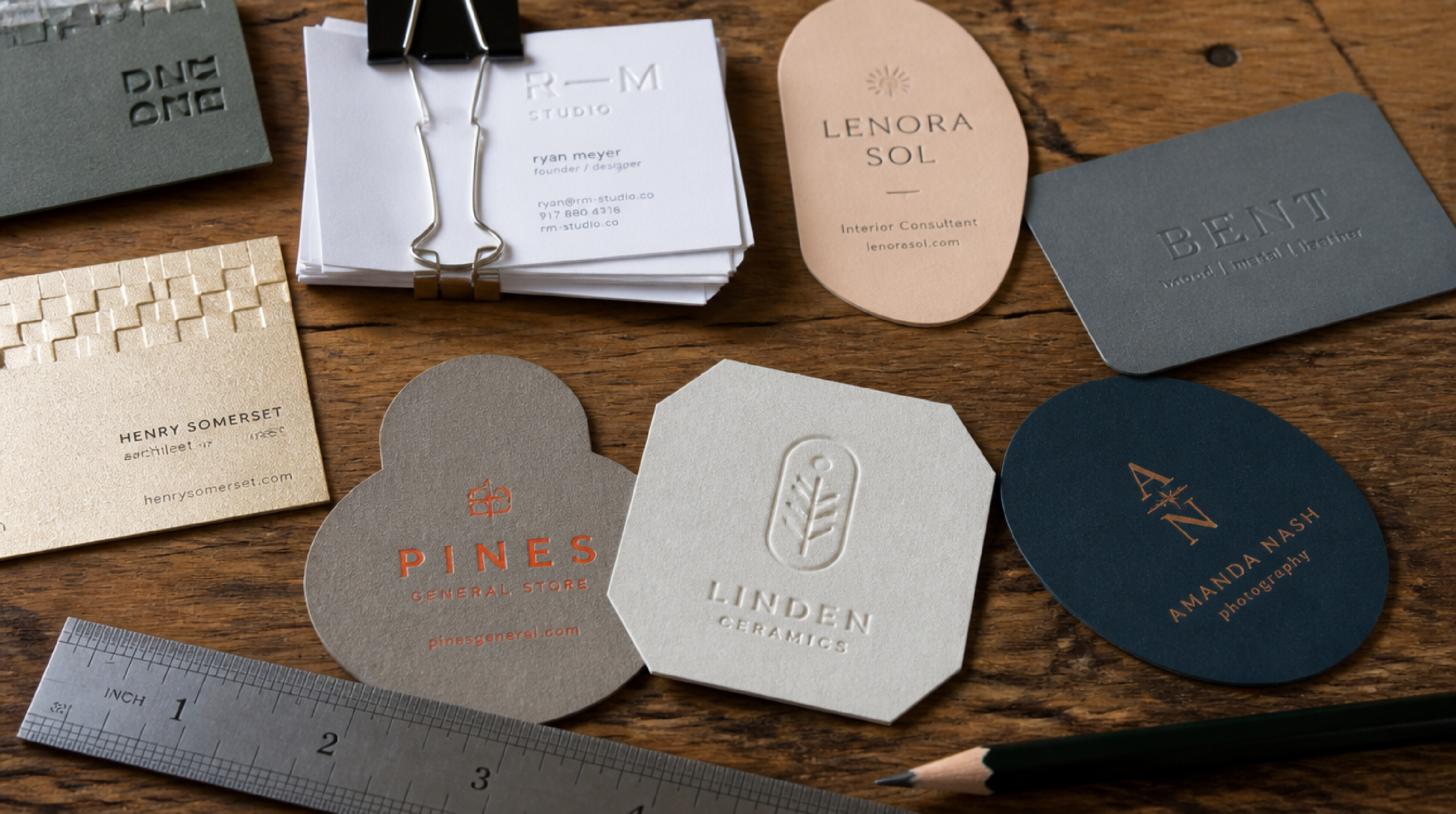 Assortment of die-cut, embossed business cards using varied shapes, typography and color, laid out on a wooden desk with a ruler